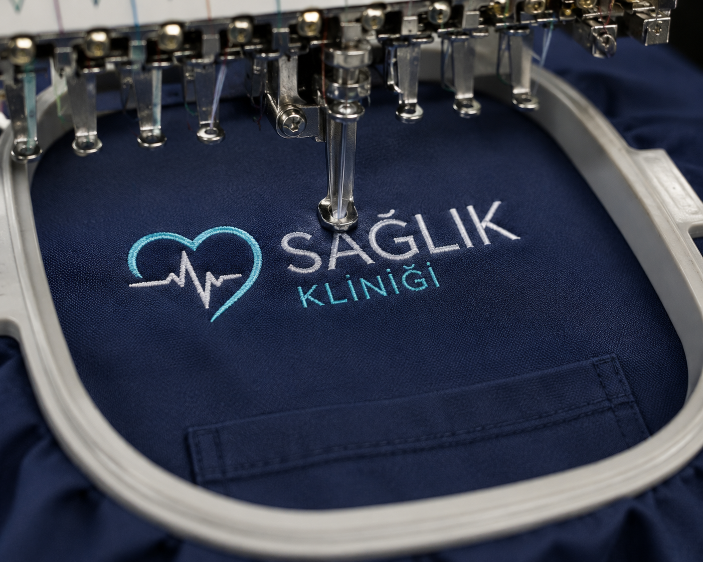 Scrubs üzerine uygulanan kurumsal logo ve isim nakışı, profesyonel nakış makineleriyle yakın çekimde gösterilmektedir. Krallar Tekstil; hastane, klinik, poliklinik, laboratuvar ve diğer sağlık kuruluşları için logolu, isim yazılı ve kuruma özel scrubs üretimi gerçekleştirmektedir.